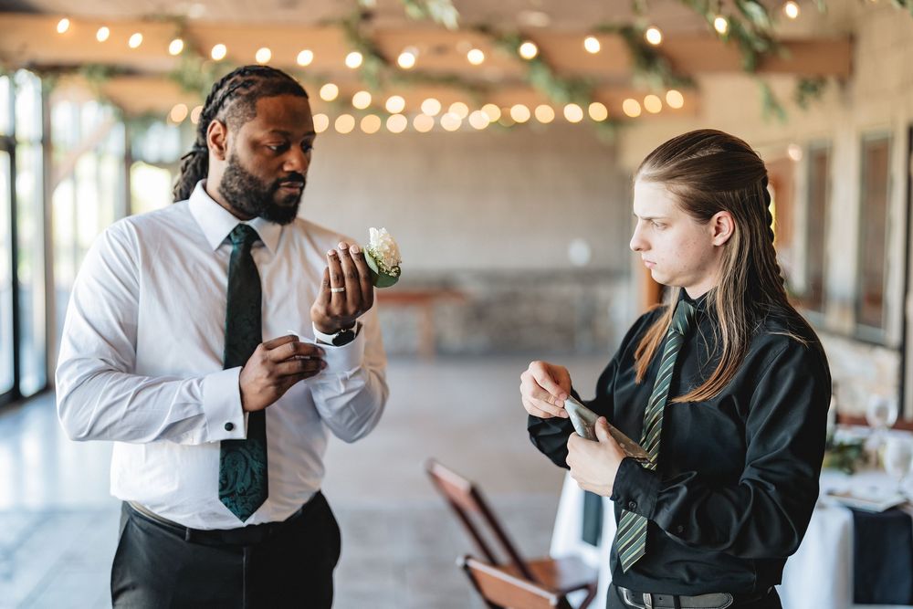 Two hospitality professionals check wine glasses in a venue decorated with string lights.
