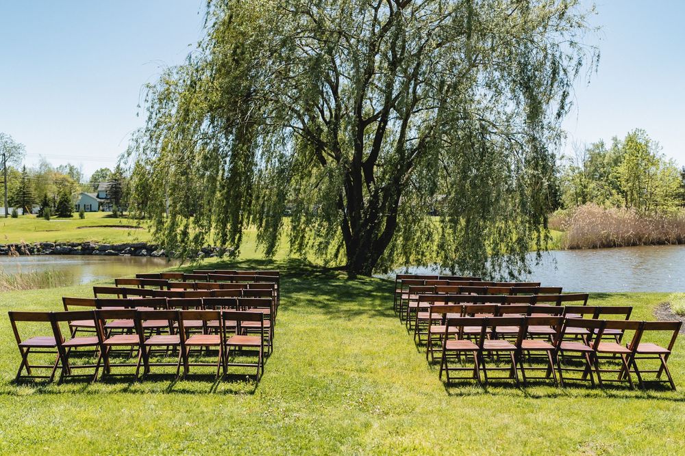 Outdoor wedding ceremony setup with wooden chairs arranged beneath a weeping willow tree by a serene pond.
