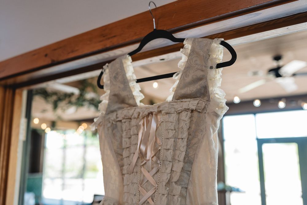 A vintage-style ivory corset wedding dress with delicate lacework and ribbons hangs on a black hanger in natural light.