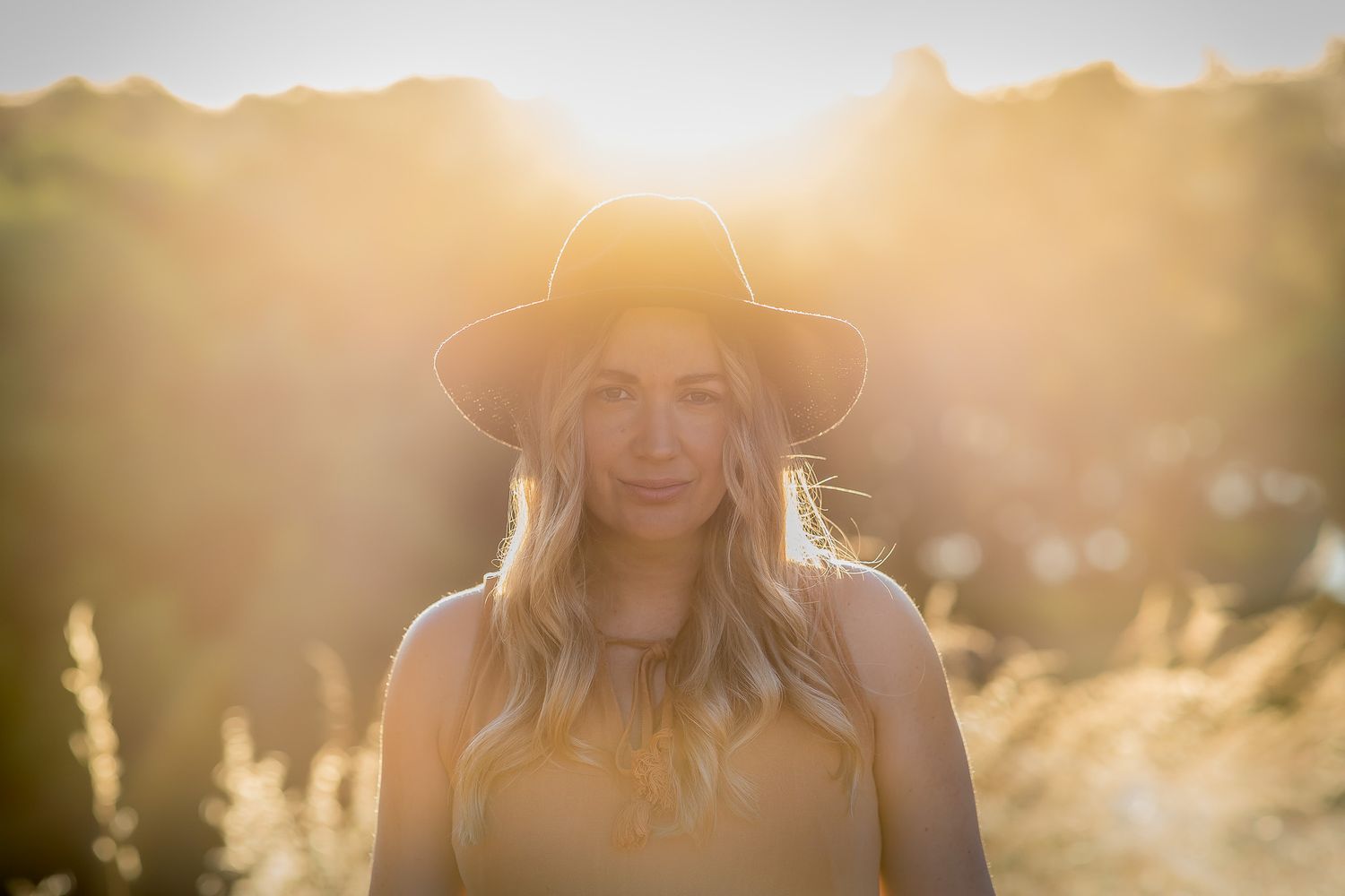 A person wearing a brown hat stands in a field during golden hour with sunlight creating a dreamy backlit effect.
