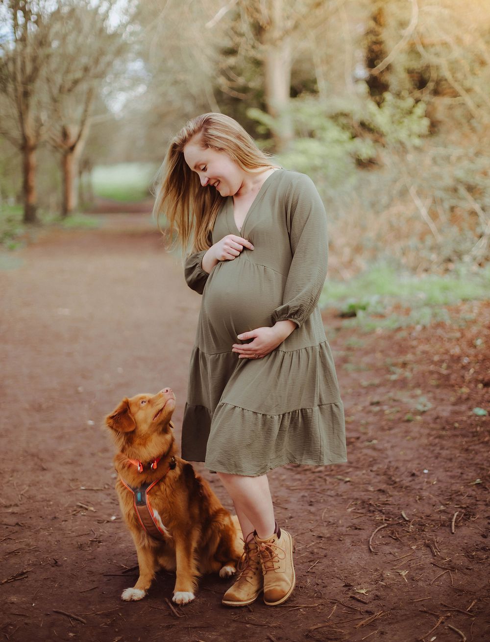 Pregnant woman in olive dress stands with golden retriever dog on autumn forest trail during maternity photoshoot.