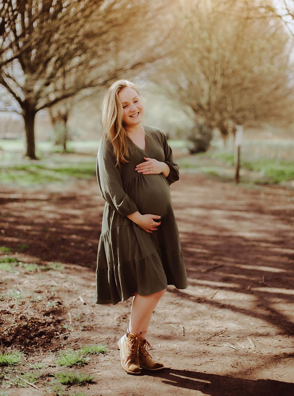 Expectant mother wearing long brown dress and boots stands on dirt path with autumn trees in background.