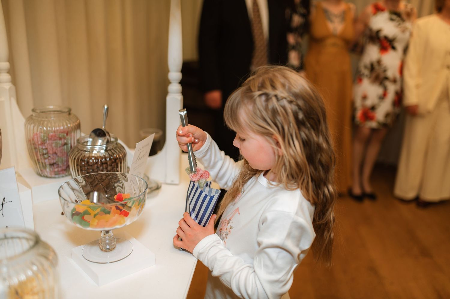 A small child in a white outfit excitedly samples treats at a dessert table with glass containers during a formal event.
