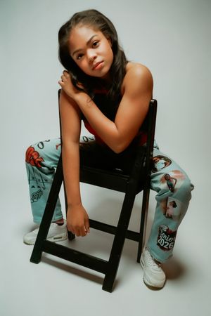 Young woman sitting on a wooden chair in printed pants and black top, posing for a studio portrait.