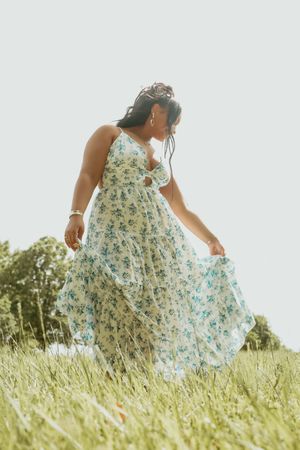Woman in floral sundress walking through a grassy field on a bright overcast day.