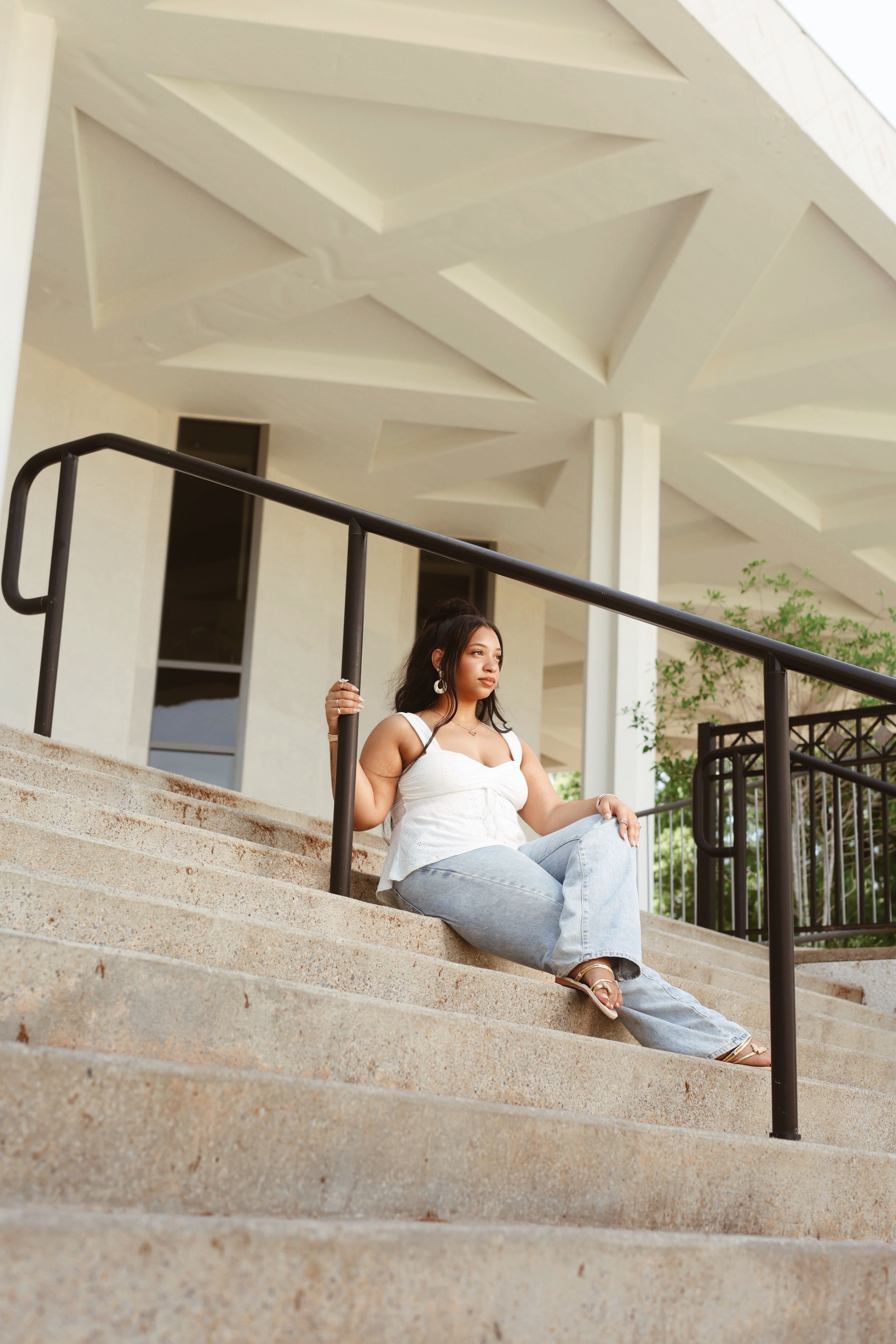 Woman in white tank and jeans sitting on outdoor staircase steps.
