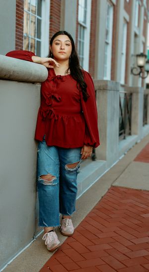 Young woman in red ruffled blouse and ripped jeans leaning against a wall in an urban setting.