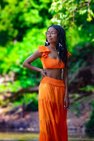 Confident young Black woman in orange two-piece outfit standing in a lush green outdoor natural setting.