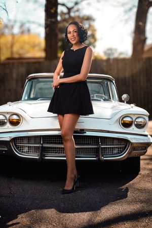 Elegant woman in black dress posing in front of a classic vintage white car outdoors.