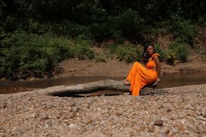 Woman in orange gown sitting on rocks by a shallow river surrounded by lush green trees.