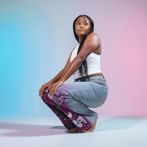 Young woman in white crop top and colorful jeans crouching against blue-pink gradient background.