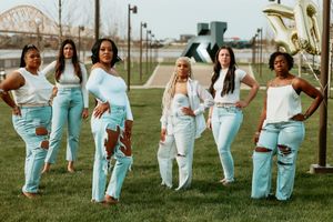 Group of six women in white tops and light blue jeans posing together outdoors near a river.