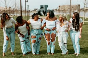 Six women in white tops and ripped light blue jeans laughing and walking together outdoors.