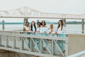 Group of six women posing on a bridge overlooking a river on a cloudy day.