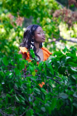 Young Black woman in orange top with white hair accessories partially framed by green leafy branches outdoors.