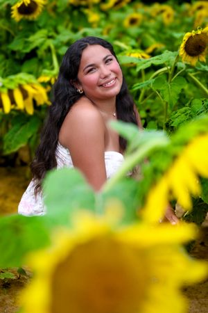 Smiling young woman with long dark hair posing among vibrant yellow sunflowers in a sunflower field.