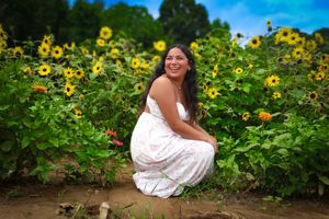 Happy young woman in white floral dress crouching beside blooming sunflowers under a bright blue sky.
