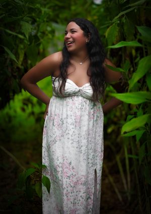 Young woman in white floral maxi dress laughing joyfully while standing among tall green tropical plants.