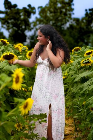 Young woman in white floral dress reaching toward sunflowers in a golden sunflower field at dusk.