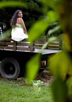Young woman in white dress sitting on flatbed truck viewed through green foliage.