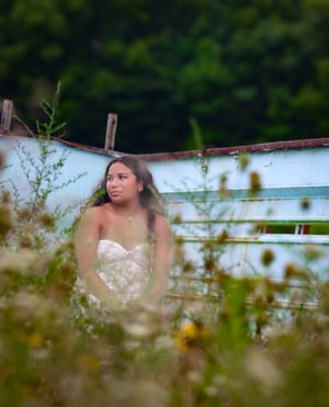 Young woman in white floral dress standing among wildflowers near a vintage turquoise boat in a lush green field.
