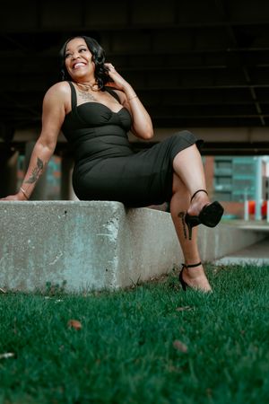 Smiling woman in black bodycon dress sits on concrete block with black heels, tattoos visible on arm.
