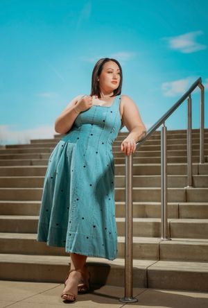Plus-size woman in light blue midi dress poses confidently on outdoor staircase under bright blue sky.