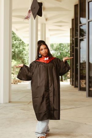 Graduating student in brown cap and gown tosses mortarboard in the air while shrugging playfully outdoors.