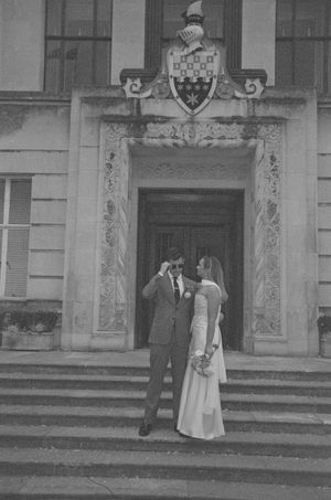 A couple stands on ornate stone steps under a decorative architectural entrance with a coat of arms above.