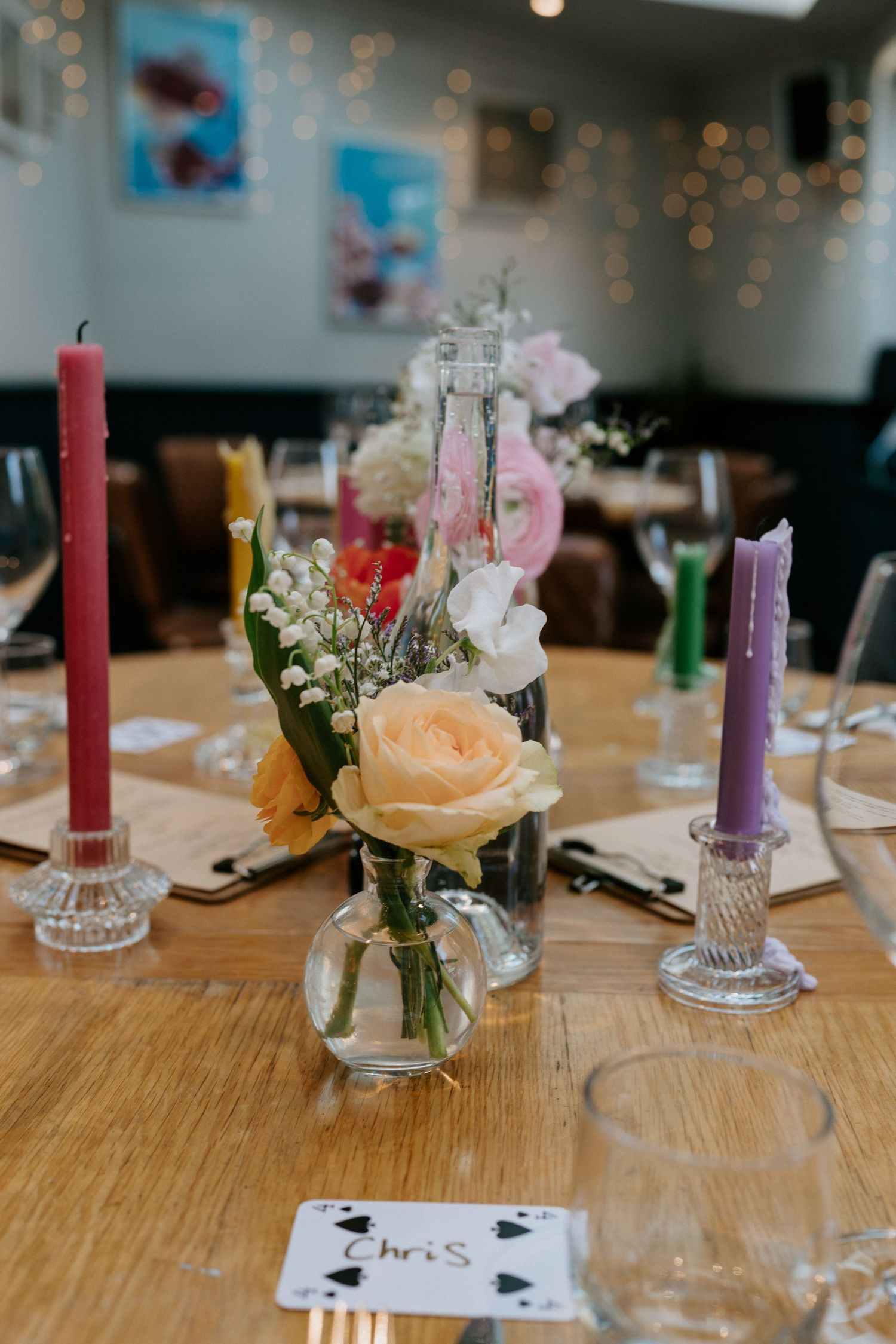 A table set with colorful candles and a mason jar centerpiece filled with peach roses and white flowers on a wooden surface.