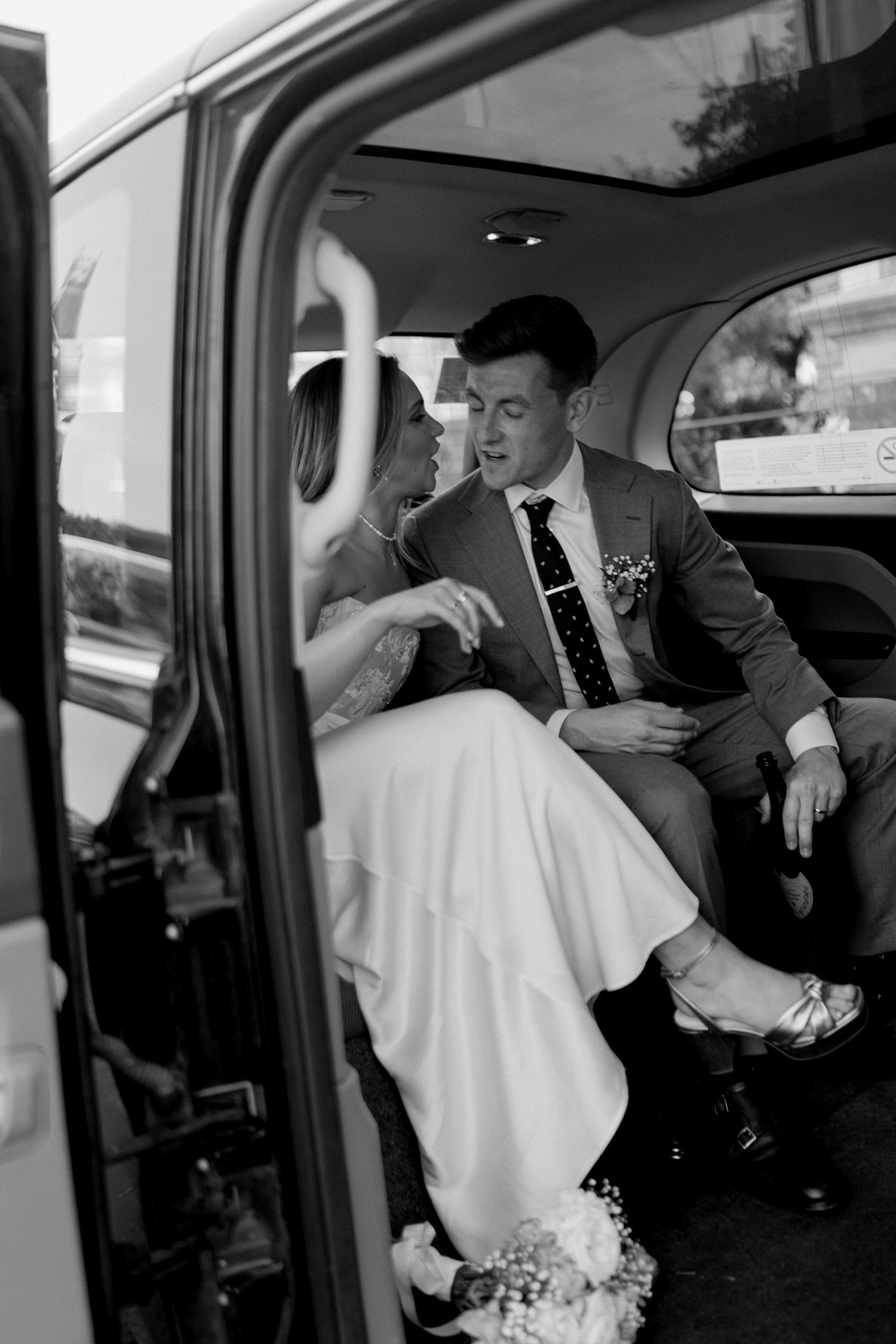 A vintage black and white wedding photo showing two people in formal attire sitting in the back seat of a classic car.