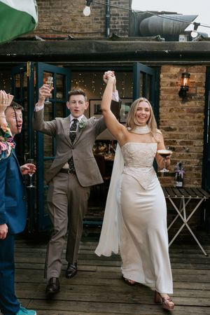 Newlyweds celebrate joyfully on a rustic wooden deck while holding champagne glasses surrounded by party lights.