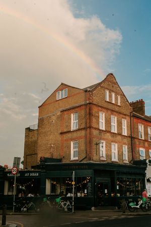 Vintage brick building with a pub on the ground floor and a vibrant rainbow arching across the cloudy sky above.