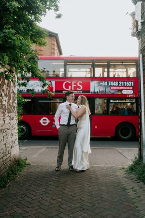 A couple in formal attire poses on a brick sidewalk with a red London double-decker bus in the background.
