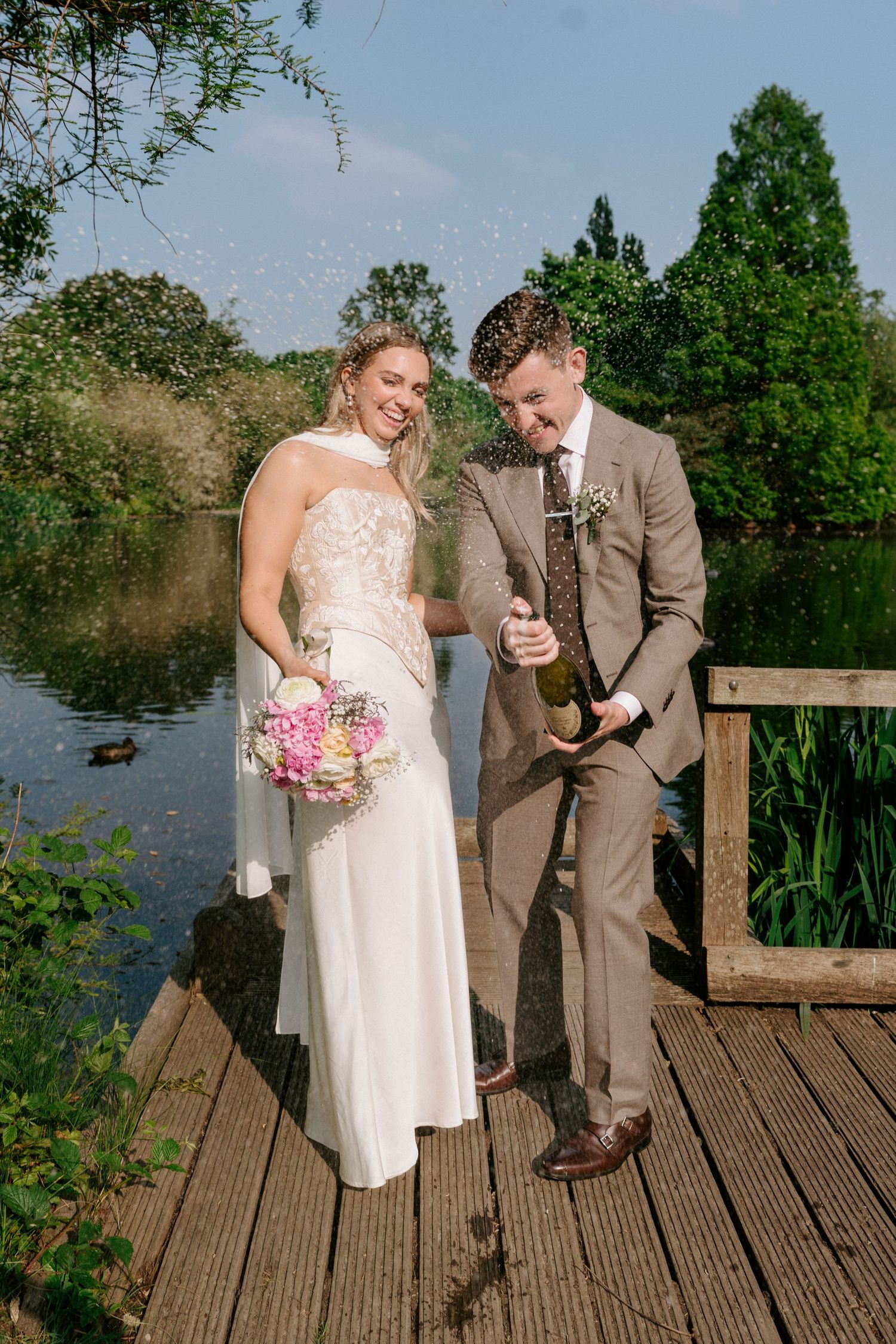 A romantic wedding couple shares a candid moment on a wooden dock by a serene pond on their special day.