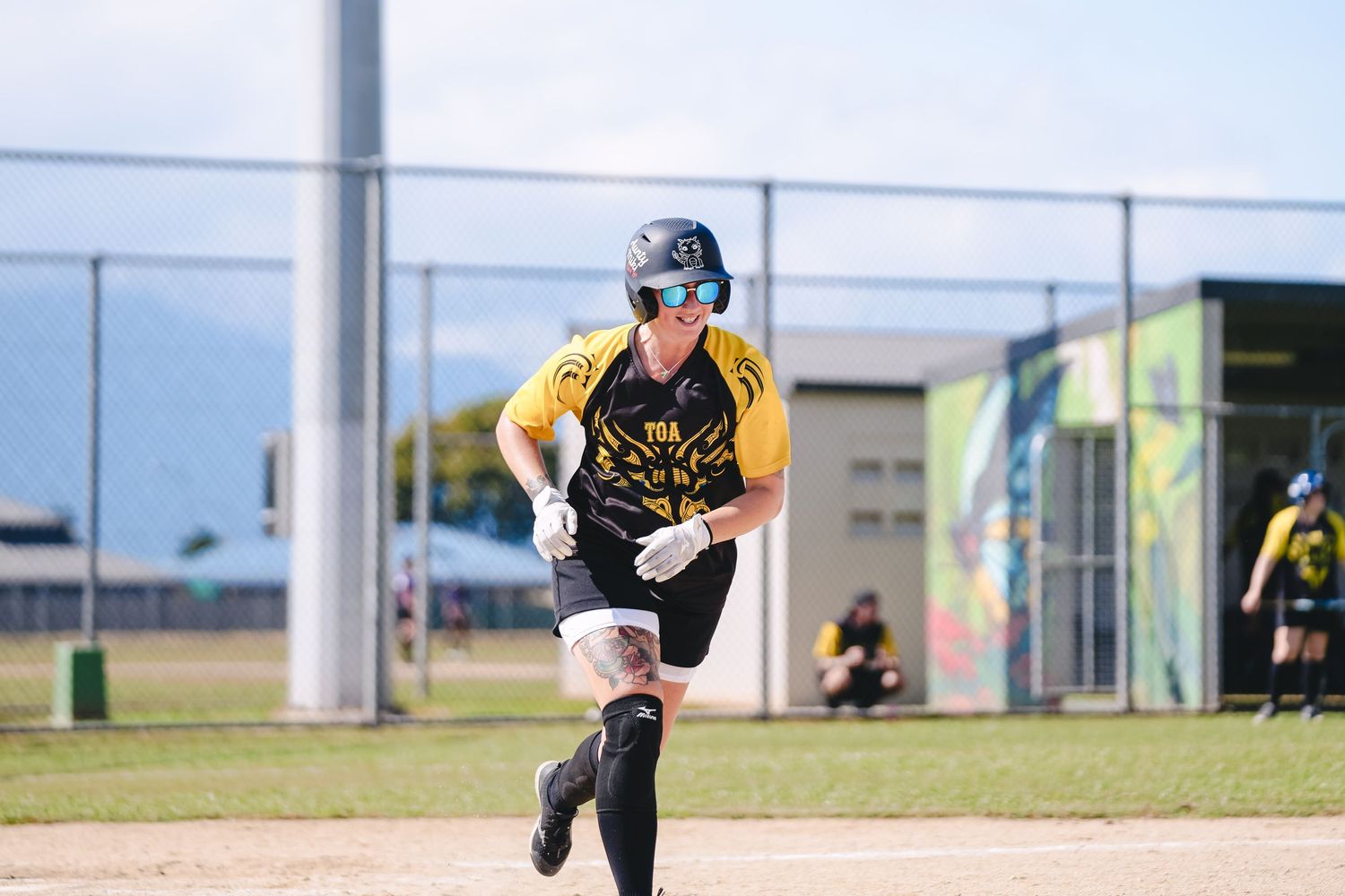 Softball player in yellow and black uniform running on baseball diamond during daytime game.