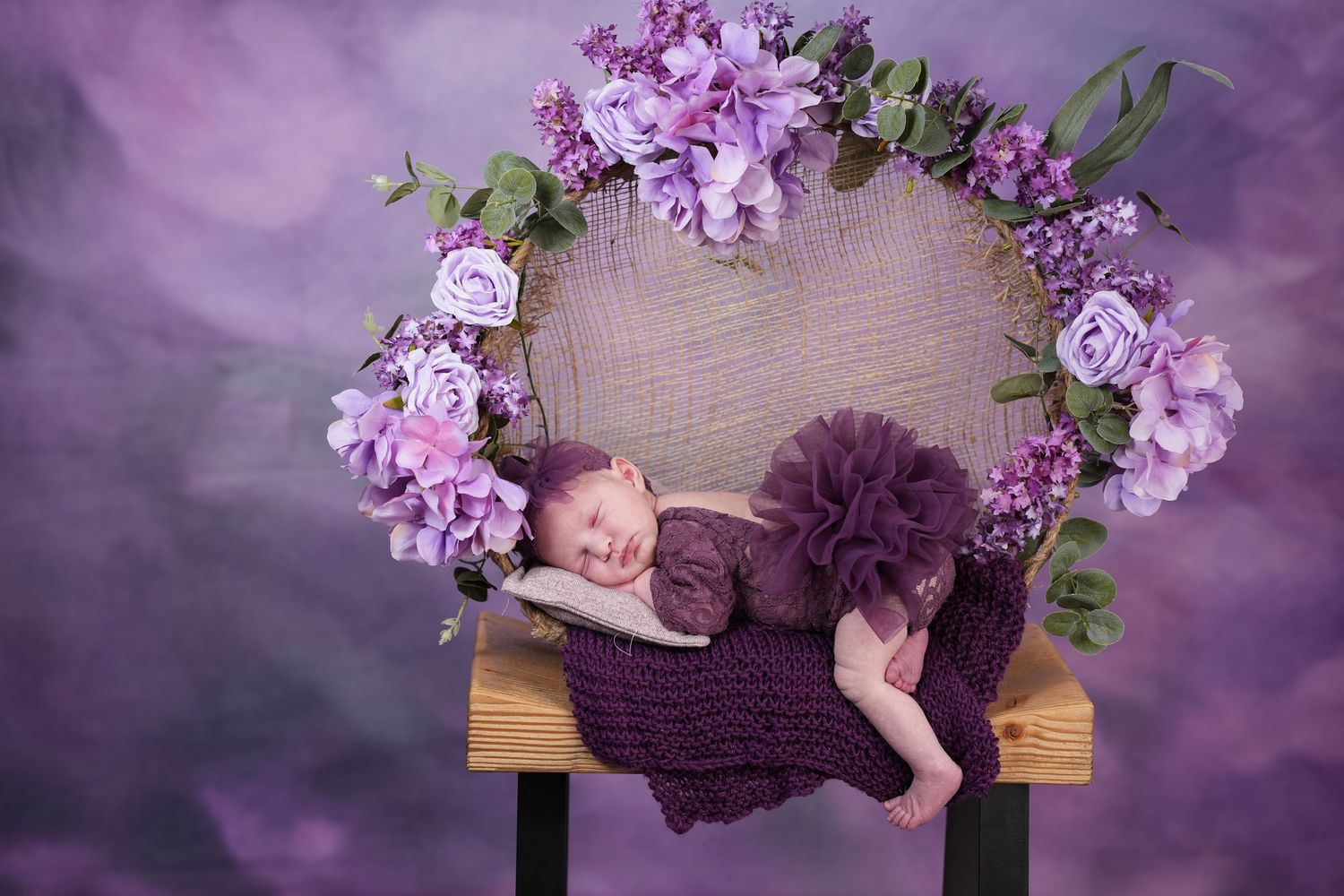 A sleeping baby lies on a wooden bench decorated with purple flowers and a tulle tutu in a dreamy purple setting.