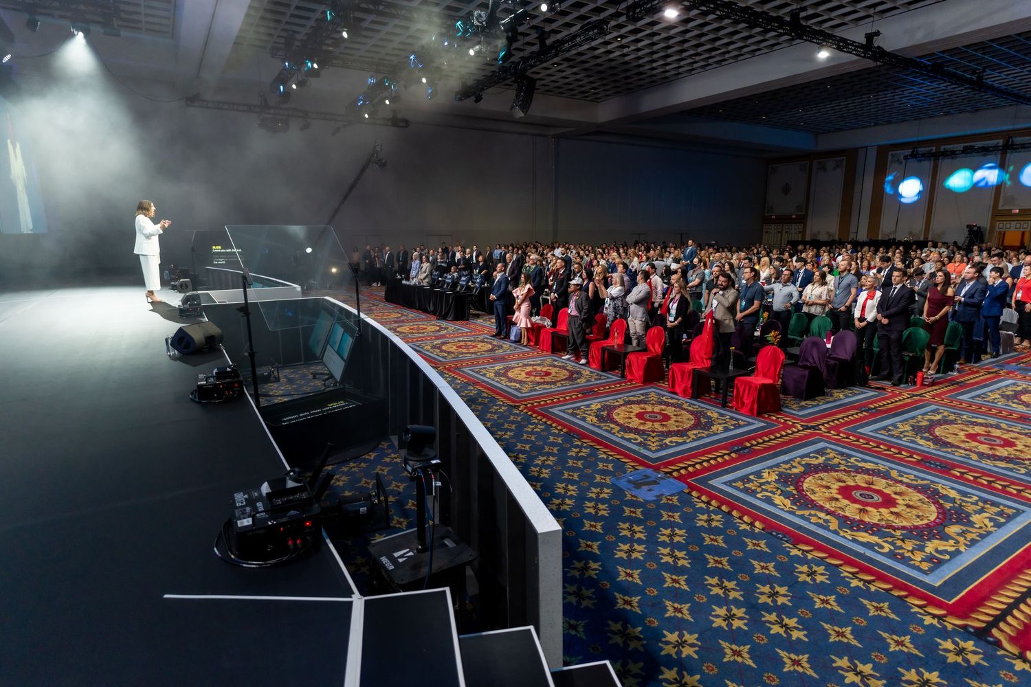 Speaker on stage addressing large audience in conference hall with decorative carpet.
