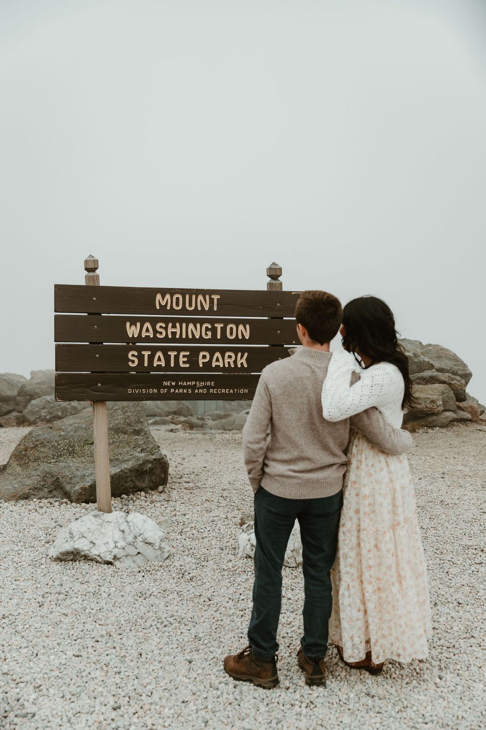 Couple standing by Mount Washington State Park sign on misty mountain summit.