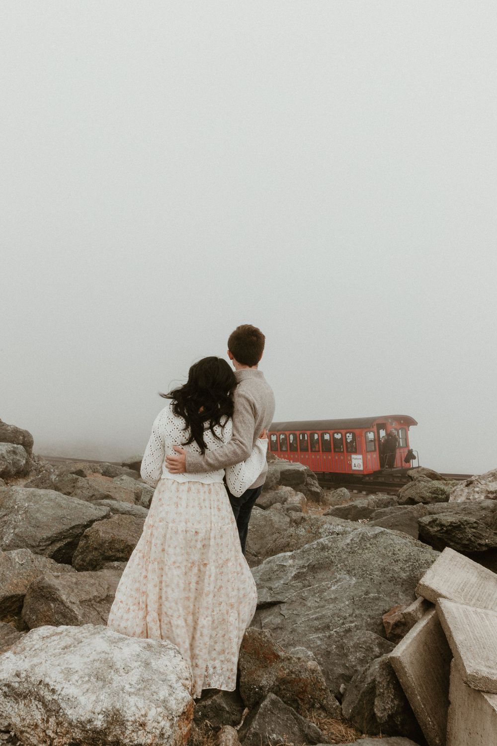 Couple embracing on rocky mountain summit with red building in background.