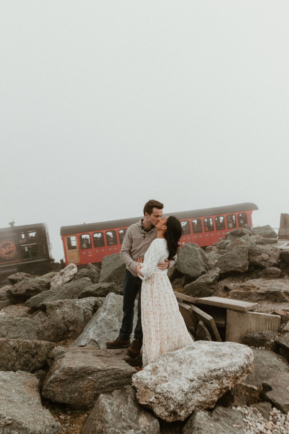A romantic couple embraces near vintage red train cars against a rocky mountain backdrop.