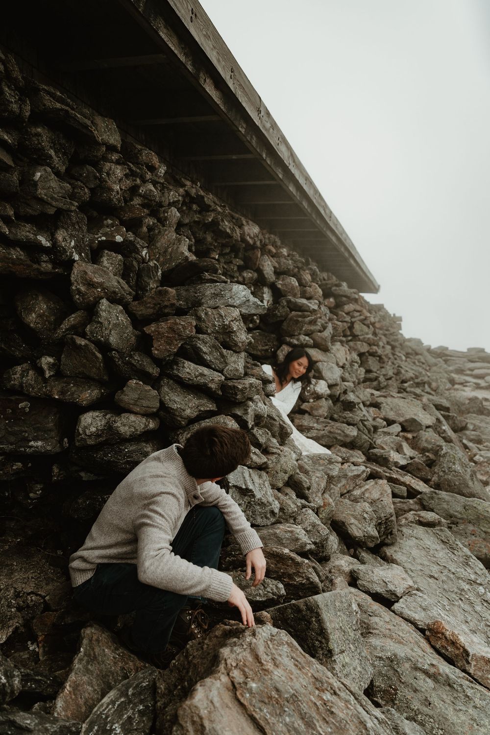 A person in a gray sweater climbs rocky terrain beneath an overhanging structure.