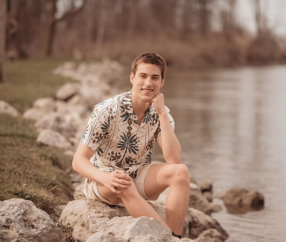 Person in floral print shirt poses casually on rocky riverbank on a warm summer day.