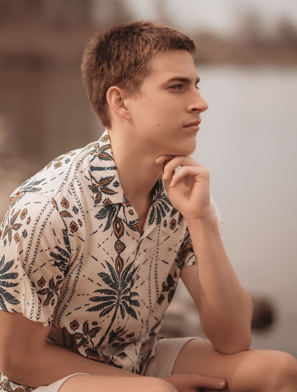 Person in a floral printed short-sleeve shirt sits thoughtfully with hand on chin against blurred background.