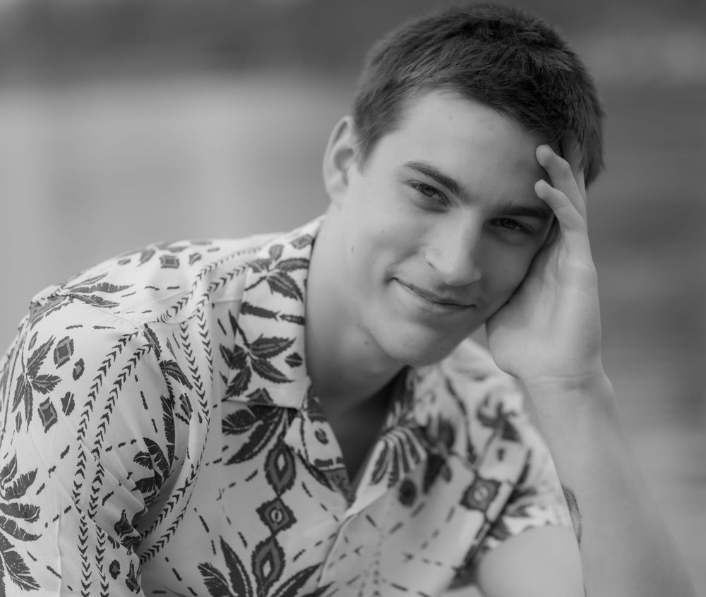 Black and white portrait photo of someone wearing a patterned shirt with leaves and floral designs leaning on their hand.