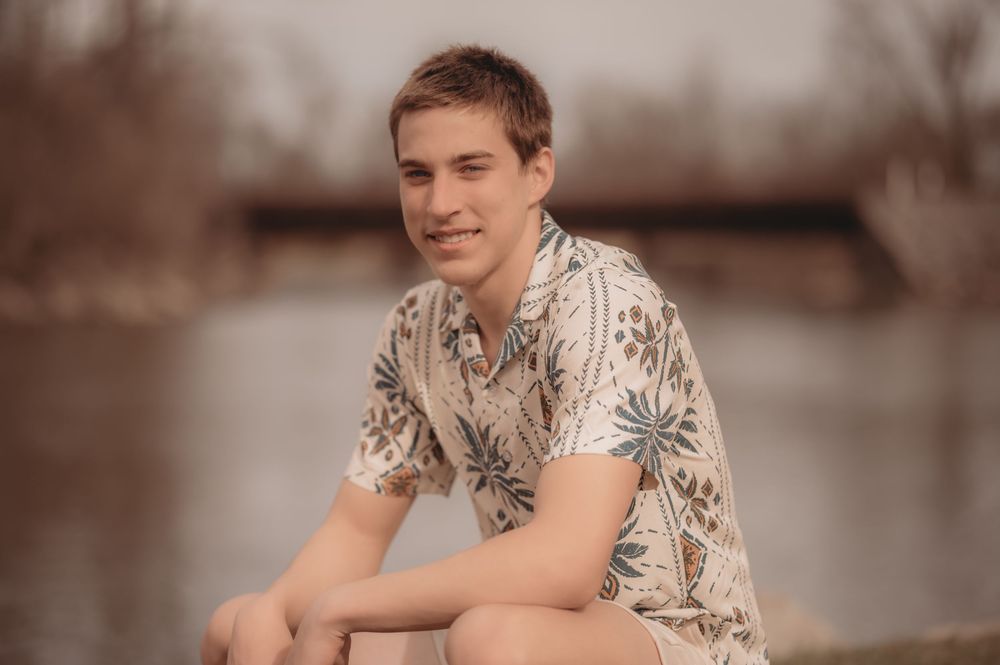 Person in a patterned short sleeve shirt sits casually outdoors near a river on an overcast day.
