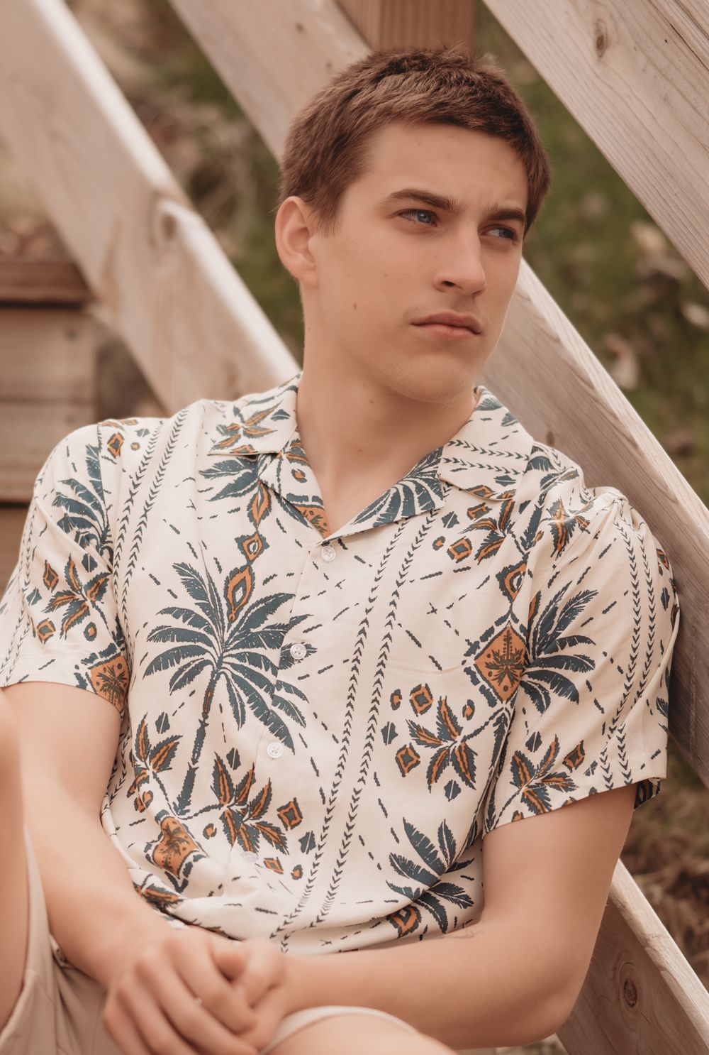 A person wearing a tropical print short-sleeve button-up shirt with palm trees and foliage sits against wooden siding.