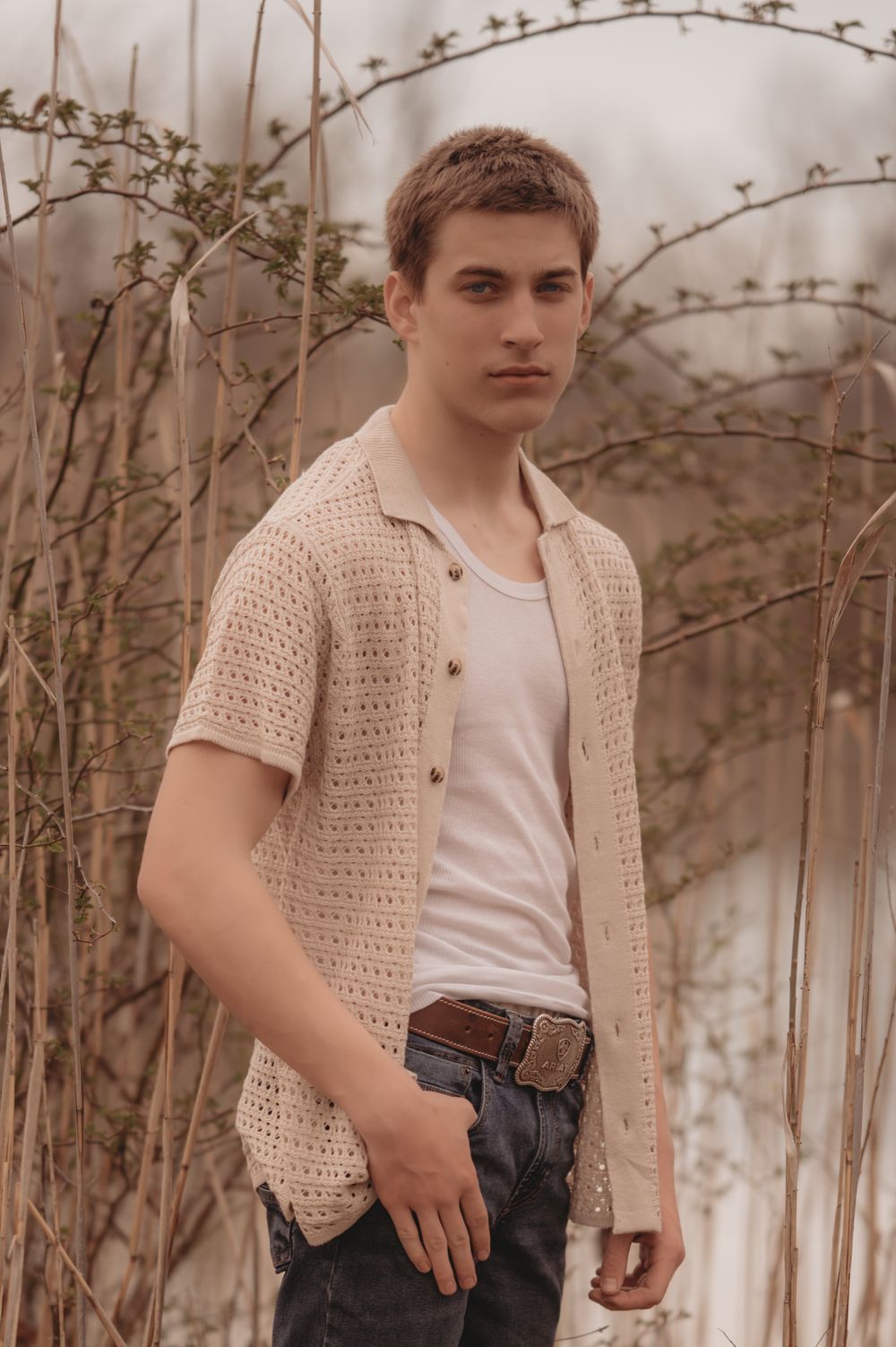 Person in cream crochet cardigan and white t-shirt poses against backdrop of barren branches in muted tones.