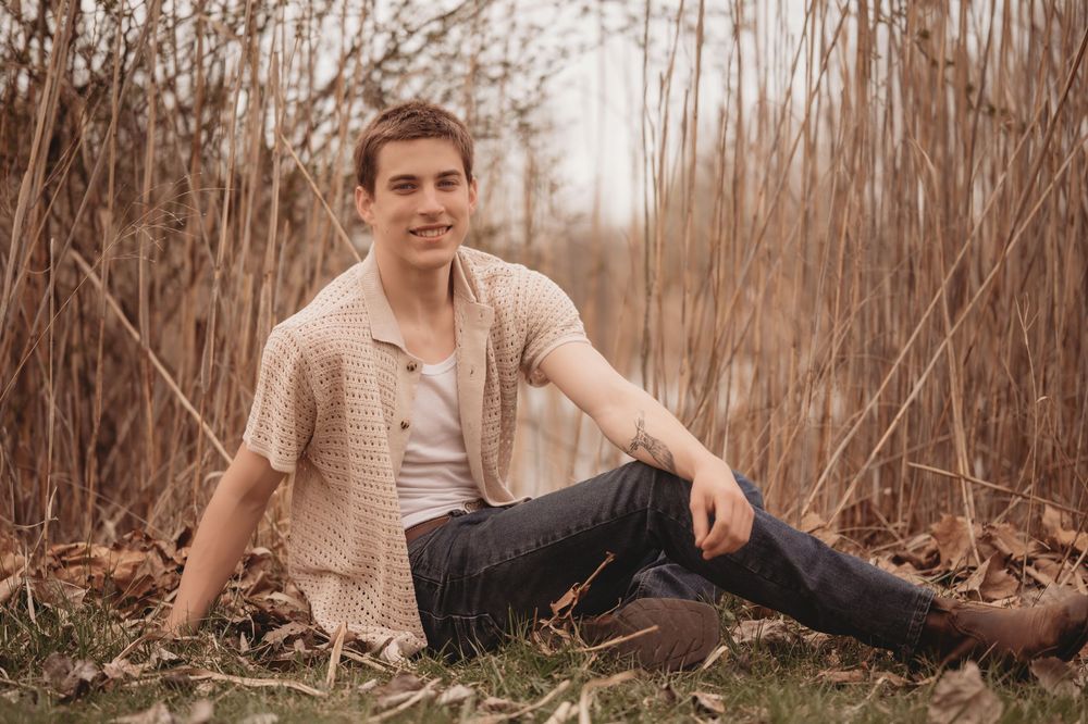 A person sits casually in field of tall brown reeds wearing a beige cardigan sweater and brown boots.