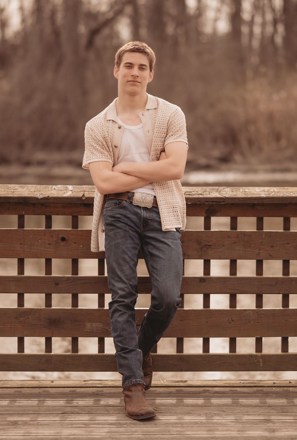 Person in casual outfit with plaid shirt and jeans poses with arms crossed on rustic wooden deck during autumn afternoon.